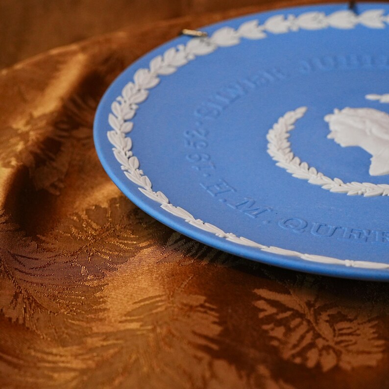 May include: A blue decorative plate with white relief detailing. The plate features a white laurel wreath border and text that reads "E.M. QUEEN 1952-SILVER JUBILEE". A white profile of a person is also visible.