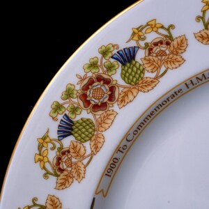 Pode incluir: Um prato de porcelana branca com borda dourada, apresentando uma borda decorativa de cardos verdes, rosas vermelhas e flores amarelas. O texto 1900. To Commemorate H.M. est&aacute; impresso em preto ao longo da borda interna do padr&atilde;o floral.