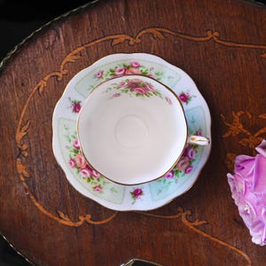 K&ouml;nnte beinhalten: Ein Vintage-Tee-Set mit Blumendesign. Die Tasse und Untertasse haben einen hellgr&uuml;nen Hintergrund mit rosa Rosen und goldenen Verzierungen. Das Set steht auf einem Holztisch mit dekorativen Intarsien.