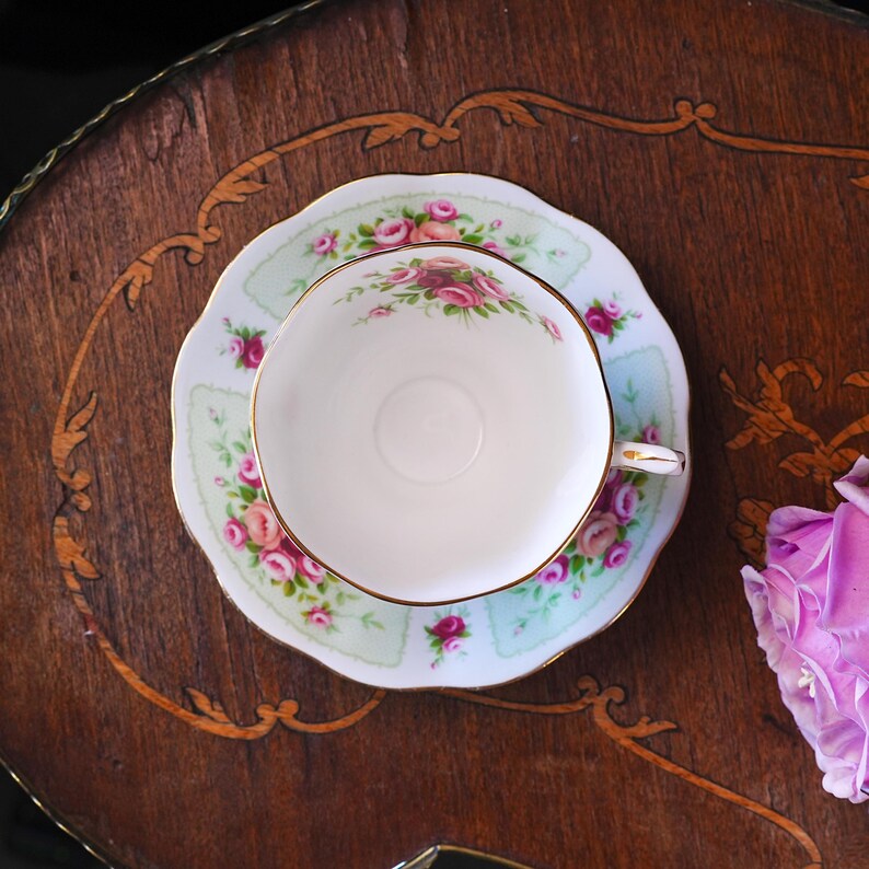K&ouml;nnte beinhalten: Ein Vintage-Tee-Set mit Blumendesign. Die Tasse und Untertasse haben einen hellgr&uuml;nen Hintergrund mit rosa Rosen und goldenen Verzierungen. Das Set steht auf einem Holztisch mit dekorativen Intarsien.