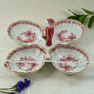 May include: A white porcelain serving dish with four petal-shaped compartments, each decorated with pink floral patterns and gold trim. A red bird figurine acts as a central handle. The dish measures approximately 25 cm across.
