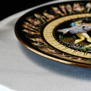 May include: A decorative black plate with a gold rim and intricate artwork. The plate features a central design with figures in motion, surrounded by a gold border and additional detailed illustrations. The plate is set against a light gray surface.