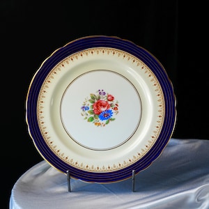 Pode incluir: Um prato decorativo de porcelana com um centro creme e uma borda azul marinho. O prato apresenta um desenho floral no centro com flores vermelhas, azuis e roxas. A borda tem um padr&atilde;o de filigrana dourada e uma borda dourada.