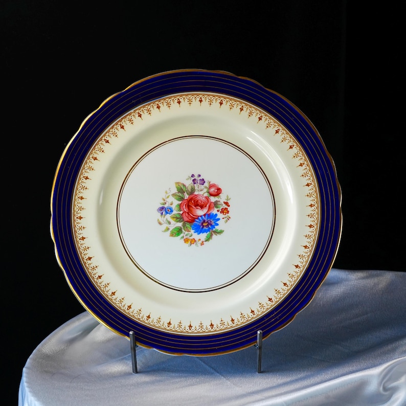 Pode incluir: Um prato decorativo de porcelana com um centro creme e uma borda azul marinho. O prato apresenta um desenho floral no centro com flores vermelhas, azuis e roxas. A borda tem um padr&atilde;o de filigrana dourada e uma borda dourada.