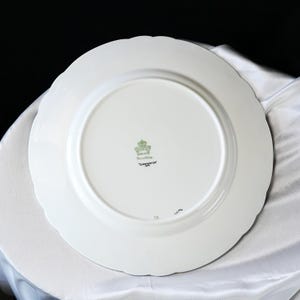 Pode incluir: Prato de cer&acirc;mica branco com borda ondulada. A parte inferior apresenta um emblema verde e dourado, as palavras "Fine China" e "Dorchester" em texto preto. O prato est&aacute; sobre uma superf&iacute;cie branca.