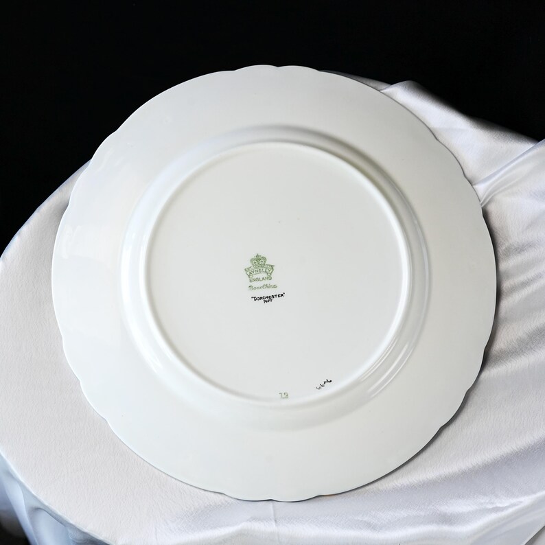 Pode incluir: Prato de cer&acirc;mica branco com borda ondulada. A parte inferior apresenta um emblema verde e dourado, as palavras "Fine China" e "Dorchester" em texto preto. O prato est&aacute; sobre uma superf&iacute;cie branca.