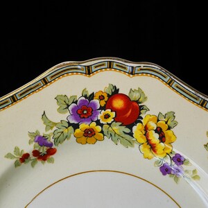 Puede incluir: Plato de cer&aacute;mica color crema con un dise&ntilde;o floral con flores amarillas, moradas y rojas, y hojas verdes. El plato tiene un borde dorado y un borde decorativo con detalles azules y dorados. El plato est&aacute; sobre un fondo negro.