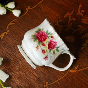 Peut inclure: Une tasse en porcelaine blanche orn&eacute;e d'un motif floral compos&eacute; de roses rose fonc&eacute;, de feuilles et de tiges vertes. La tasse pr&eacute;sente une base et un bord festonn&eacute;s, avec une bordure m&eacute;tallique dor&eacute;e sur le dessus et une anse incurv&eacute;e.