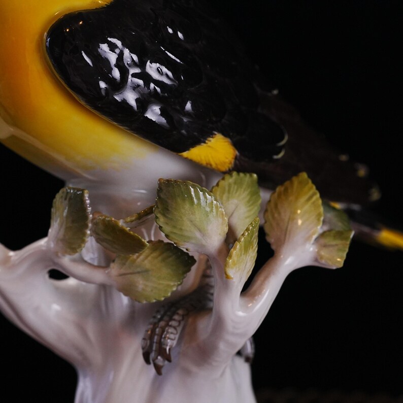 May include: A detailed porcelain figurine of a bird with yellow, black, and white plumage perched on a white branch with green leaves. The bird's feathers are intricately detailed, and the leaves have a glossy finish.