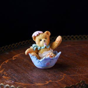 May include: A charming figurine of a teddy bear emerging from a blue eggshell. The bear is light brown with a pink sun hat and a blue bib. The eggshell is decorated with purple and pink accents, creating a whimsical Easter theme.