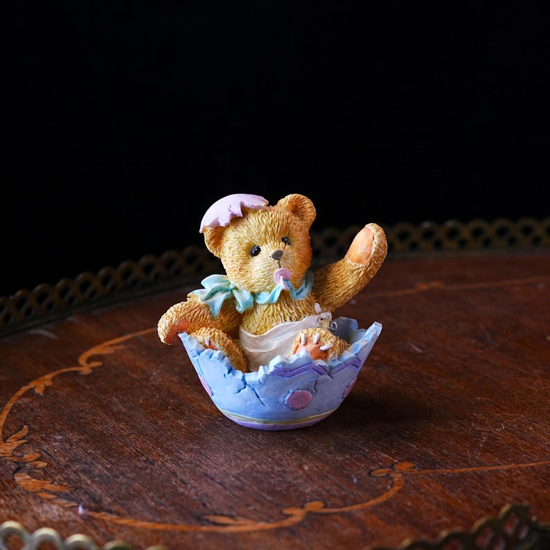 May include: A charming figurine of a teddy bear emerging from a blue eggshell. The bear is light brown with a pink sun hat and a blue bib. The eggshell is decorated with purple and pink accents, creating a whimsical Easter theme.