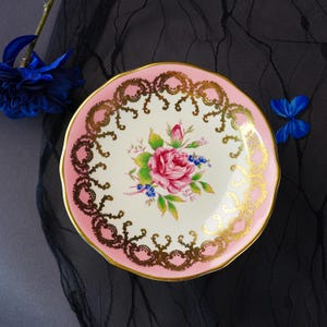 Puede incluir: Un platillo redondo de cer&aacute;mica con un centro de color crema que presenta un motivo floral de una rosa rosa y hojas verdes. El borde est&aacute; pintado de rosa y decorado con un intrincado patr&oacute;n de filigrana dorada. El borde tiene un acabado festoneado.