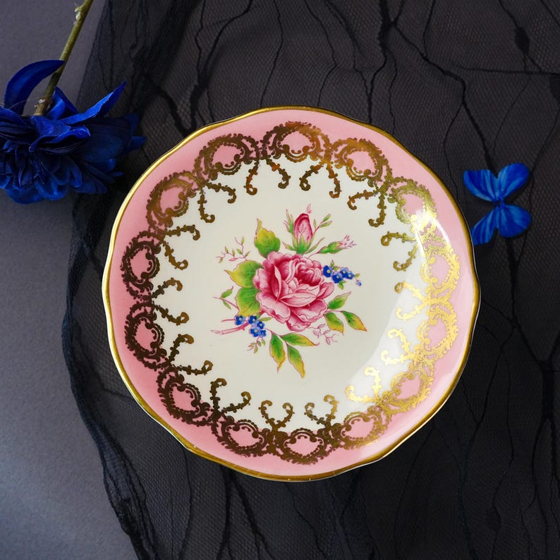 Puede incluir: Un platillo redondo de cer&aacute;mica con un centro de color crema que presenta un motivo floral de una rosa rosa y hojas verdes. El borde est&aacute; pintado de rosa y decorado con un intrincado patr&oacute;n de filigrana dorada. El borde tiene un acabado festoneado.