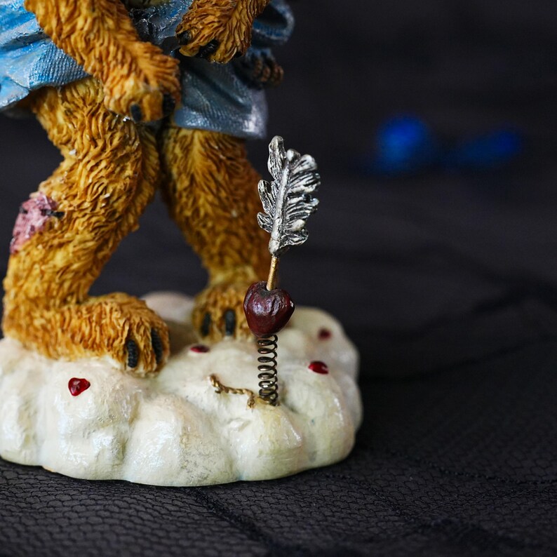 Puede incluir: Una figura de oso marr&oacute;n texturizado con una prenda azul sobre una base blanca en forma de nube. La base presenta peque&ntilde;os corazones rojos y una flecha plateada con un coraz&oacute;n rojo oscuro unido a un resorte met&aacute;lico que sobresale.