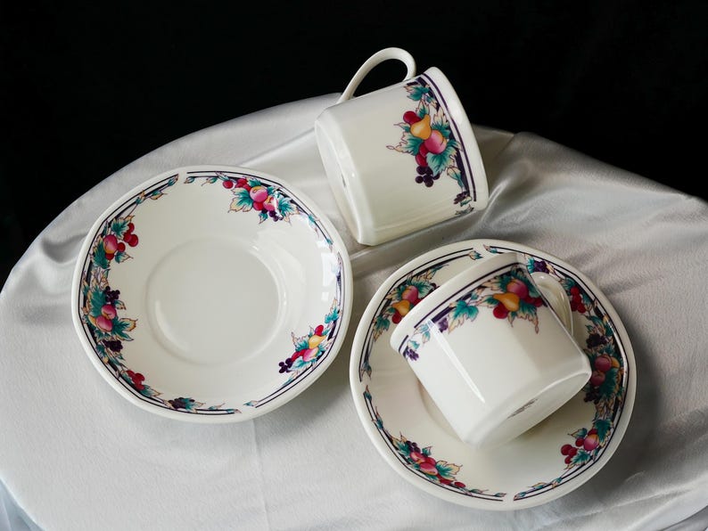 Może przedstawiać: Biały porcelanowy zestaw do herbaty z motywem owoc&oacute;w i kwiat&oacute;w. Zestaw zawiera dwie filiżanki do herbaty z uchwytami i dwa spodki. Krawędzie filiżanek i spodk&oacute;w zdobi obramowanie z owoc&oacute;w i liści.