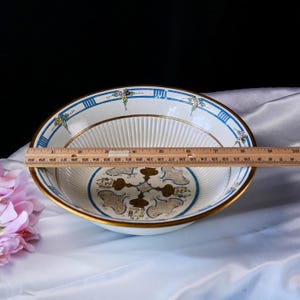 May include: An antique white ceramic bowl with a gold rim and a blue and gold decorative border. The bowl has a fluted interior and a central design with gold and floral accents. The bowl measures approximately 30 cm in diameter.