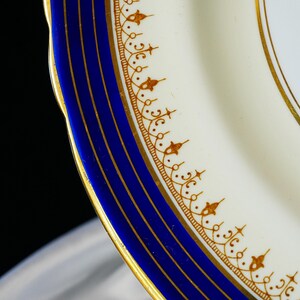 Pode incluir: Close-up de um prato de porcelana branca com uma borda azul e detalhes dourados. O prato apresenta um padr&atilde;o de filigrana dourada detalhado ao longo da borda e listras douradas dentro da borda azul. O prato tem uma borda ondulada.