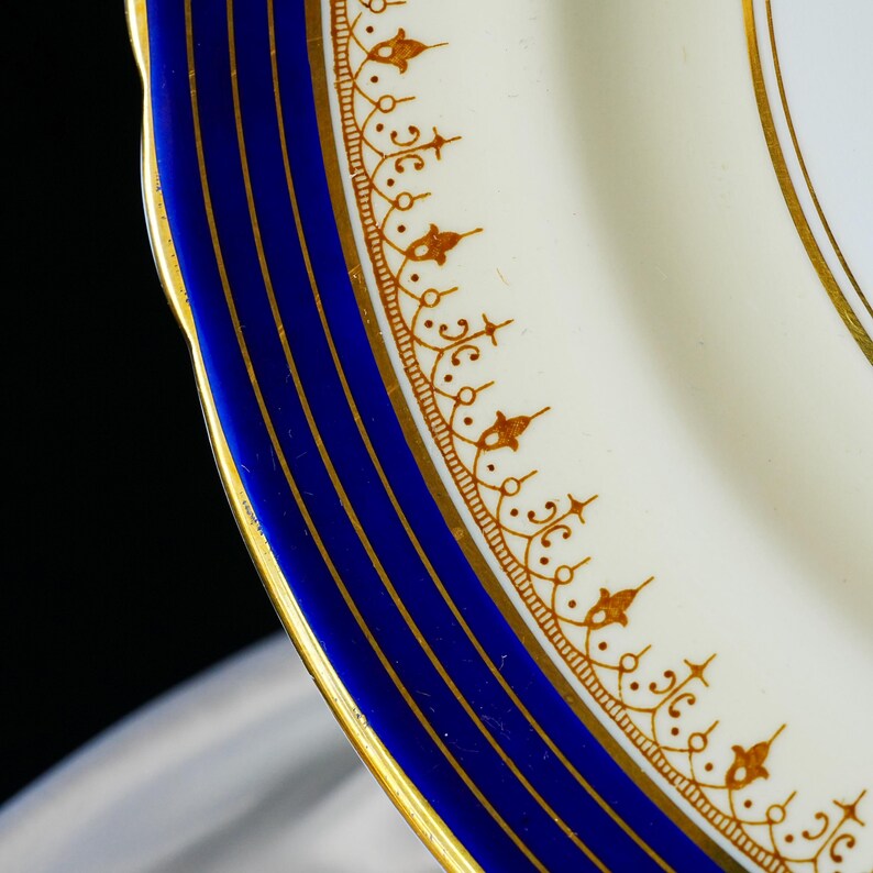 Pode incluir: Close-up de um prato de porcelana branca com uma borda azul e detalhes dourados. O prato apresenta um padr&atilde;o de filigrana dourada detalhado ao longo da borda e listras douradas dentro da borda azul. O prato tem uma borda ondulada.