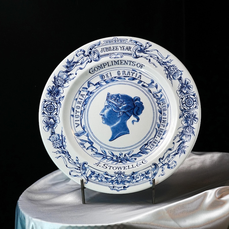 May include: A white and blue commemorative plate featuring a profile of Queen Victoria. The plate has a floral border and text that reads "Jubilee Year" and "Compliments of Dei Gratia". The plate is supported by a metal stand.