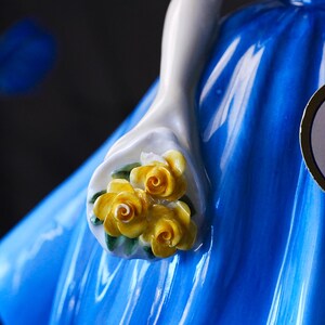 May include: A glossy ceramic figurine featuring a white arm holding a small bouquet of three yellow roses with green leaves. The figurine is wearing a vibrant, solid blue dress with a smooth, reflective finish and visible vertical folds.