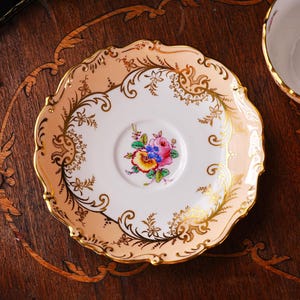 Op de afbeelding: Een vintage theekopschotel met een bloemenontwerp. De schotel heeft een geschulpte rand met een perzikkleurige rand en gouden accenten. Het midden heeft een boeket kleurrijke bloemen. Een bijpassende theekop is gedeeltelijk zichtbaar.