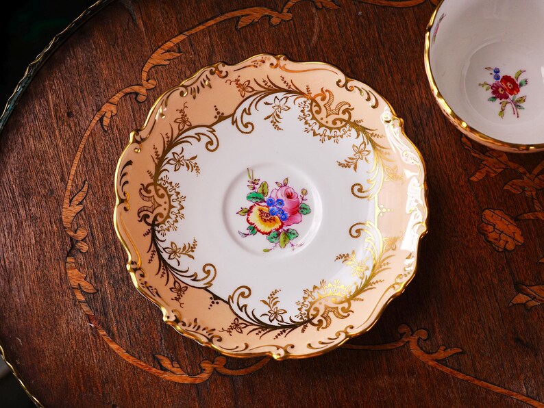 Op de afbeelding: Een vintage theekopschotel met een bloemenontwerp. De schotel heeft een geschulpte rand met een perzikkleurige rand en gouden accenten. Het midden heeft een boeket kleurrijke bloemen. Een bijpassende theekop is gedeeltelijk zichtbaar.