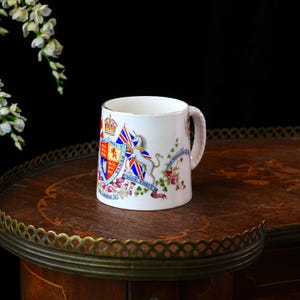 Op de afbeelding: Witte keramische mok met een wapen met kroon, vlaggen en een eenhoorn. De mok heeft een gouden rand en een gedraaide handgreep. Bloemen- en lintdetails zijn ook aanwezig. Een vintage verzamelobject.
