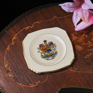 May include: A square cream-colored ceramic plate with rounded corners and a gold-trimmed edge. The center features a colorful coat of arms of Canada, depicting a lion, a unicorn, a shield, and a crown, with the word CANADA printed below it.