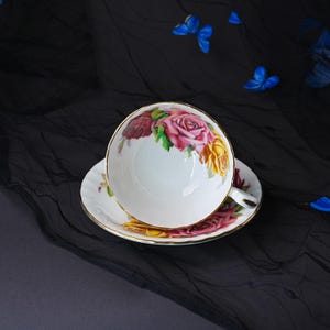 Peut inclure: Un ensemble tasse et soucoupe en porcelaine blanche orn&eacute; d'un motif floral aux roses roses, rouges et jaunes. Le bord de la tasse et de la soucoupe est soulign&eacute; d'un fin liser&eacute; dor&eacute;, et la tasse est dot&eacute;e d'une petite anse sur le c&ocirc;t&eacute;.