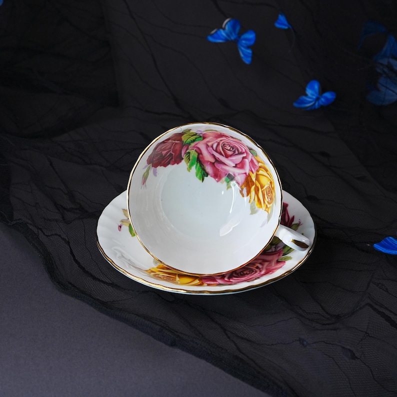 Peut inclure: Un ensemble tasse et soucoupe en porcelaine blanche orn&eacute; d'un motif floral aux roses roses, rouges et jaunes. Le bord de la tasse et de la soucoupe est soulign&eacute; d'un fin liser&eacute; dor&eacute;, et la tasse est dot&eacute;e d'une petite anse sur le c&ocirc;t&eacute;.