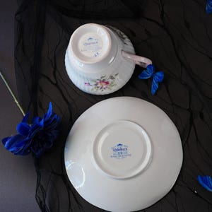 May include: A white porcelain teacup and saucer set featuring a floral pattern and a handle. The underside of both pieces displays the blue Queens Rosina China Co Ltd stamp. A blue butterfly and a blue flower are positioned near the set.