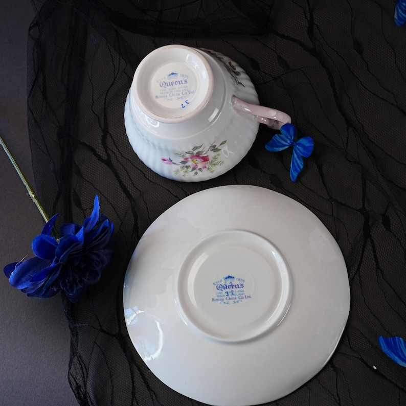 May include: A white porcelain teacup and saucer set featuring a floral pattern and a handle. The underside of both pieces displays the blue Queens Rosina China Co Ltd stamp. A blue butterfly and a blue flower are positioned near the set.