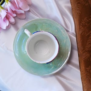 May include: A white teacup with a blue and pink rim sits on a matching green saucer. The teacup has a white handle. The set is placed on a white and brown fabric background, with pink flowers in the upper left corner.
