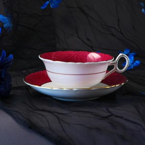 K&ouml;nnte beinhalten: Ein wei&szlig;es Teetassen- und Untertassenset mit tiefrotem Innenraum und Rand. Die Tasse hat einen gewellten Rand, eine d&uuml;nne rote horizontale Linie an der Au&szlig;enseite und einen verzierten Henkel. Die Untertasse hat einen passenden roten Rand mit Gold.