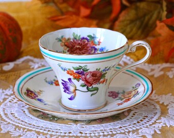 May include: A white teacup and saucer set with floral designs in shades of purple, red, orange, and blue. The cup and saucer have a turquoise stripe and gold trim. The set is placed on a white lace doily.