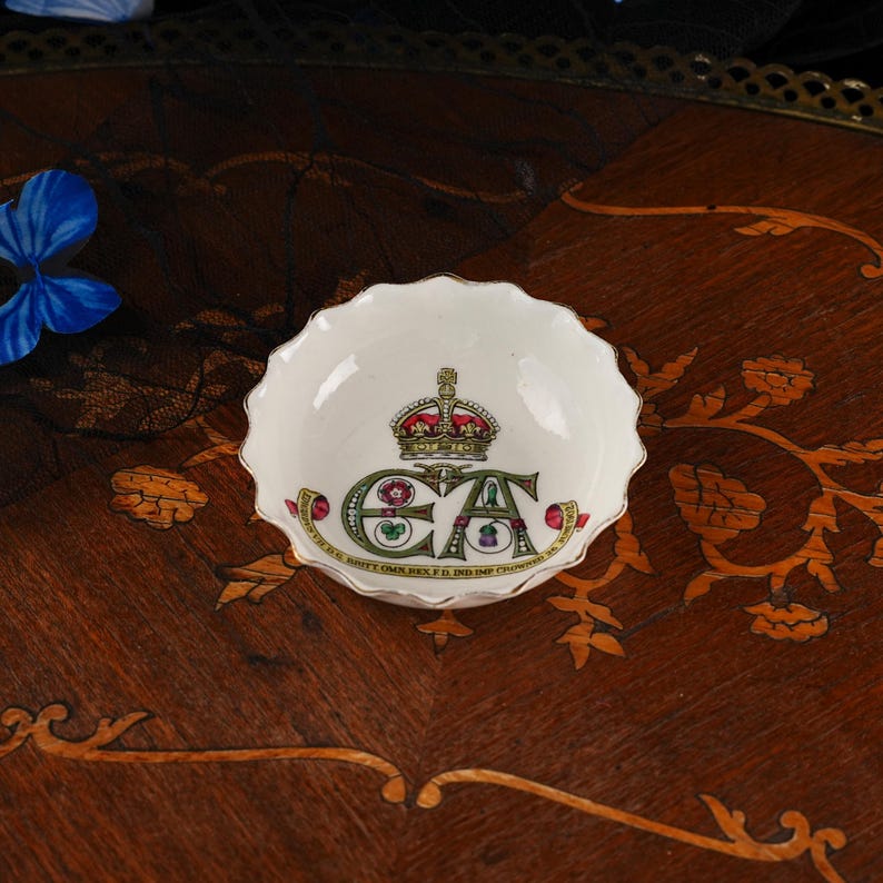 Op de afbeelding: Een kleine, ronde, witte porseleinen schaal met een geschulpte rand. Het midden toont een kleurrijke illustratie van een koninklijke kroon boven de letters EA, omringd door bloemmotieven en de tekst EDWARD VII D.G. BRITT OMN. REX F.D. IND. IMP.