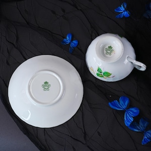 Peut inclure: Un ensemble tasse et soucoupe en porcelaine blanche orn&eacute; de motifs de feuilles et de fleurs vertes. Le dessous des deux pi&egrave;ces pr&eacute;sente un tampon vert Aynsley England avec un logo en forme de couronne. La tasse poss&egrave;de une anse courb&eacute;e.