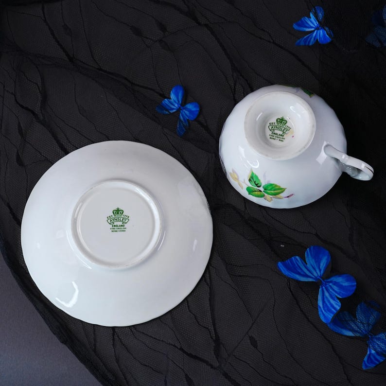 Peut inclure: Un ensemble tasse et soucoupe en porcelaine blanche orn&eacute; de motifs de feuilles et de fleurs vertes. Le dessous des deux pi&egrave;ces pr&eacute;sente un tampon vert Aynsley England avec un logo en forme de couronne. La tasse poss&egrave;de une anse courb&eacute;e.