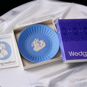 May include: A light blue Wedgwood jasperware plate with a white relief depicting figures, housed in a box with purple and white branding. Beside it, a white booklet features a blue circular portrait and the text Wedgwood and tradition.