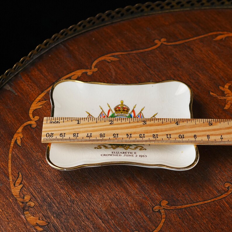 Puede incluir: Un plato peque&ntilde;o rectangular de cer&aacute;mica blanca con borde dorado, que presenta un escudo real colorido y el texto ELIZABETH II CROWNED JUNE 2 1953. Una regla de madera est&aacute; colocada sobre el plato, mostrando un ancho de unos 12,7 cm.