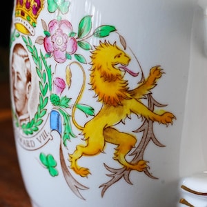 Może przedstawiać: Biały ceramiczny kubek z kolorowym nadrukiem przedstawiającym kr&oacute;la Edwarda VIII. Wz&oacute;r zawiera ż&oacute;łtego lwa, koronę, r&oacute;żowe r&oacute;że, zielone koniczyny oraz portret monarchy z napisem Edward VIII wydrukowanym pod obrazem.