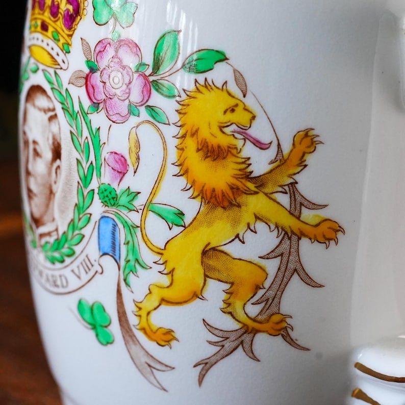 Może przedstawiać: Biały ceramiczny kubek z kolorowym nadrukiem przedstawiającym kr&oacute;la Edwarda VIII. Wz&oacute;r zawiera ż&oacute;łtego lwa, koronę, r&oacute;żowe r&oacute;że, zielone koniczyny oraz portret monarchy z napisem Edward VIII wydrukowanym pod obrazem.