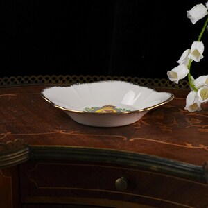 May include: A white porcelain shallow bowl with a scalloped edge and a gold-trimmed rim. The center of the bowl features a circular emblem with a crown, a portrait, and green leaf accents. The bowl has a smooth, glossy finish and curved sides.