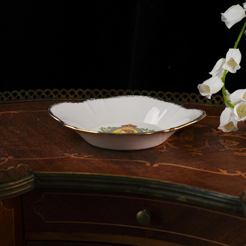 May include: A white porcelain shallow bowl with a scalloped edge and a gold-trimmed rim. The center of the bowl features a circular emblem with a crown, a portrait, and green leaf accents. The bowl has a smooth, glossy finish and curved sides.