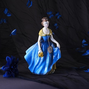 May include: A porcelain figurine of a person wearing a vibrant blue gown with a yellow shawl and holding a bouquet of yellow flowers. A gold-toned chain hangs around the neck, featuring a circular tag with the text Royal Doulton Fine Bone China.