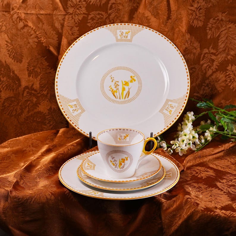 Op de afbeelding: Een wit porseleinen theeservies met een gele bloemenprint. De set bevat een groot bord, een theekopje, een schotel en een kleiner bord. Het kopje heeft een gele oor. De borden en de kop hebben een gouden rand.