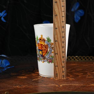 Puede incluir: Un vaso de cer&aacute;mica blanca con un escudo real colorido con una corona y el texto 'Honi Soit Qui Mal Y Pense' y 'AD 1911'. Una regla de madera est&aacute; colocada contra el lado, mostrando que el recipiente mide aproximadamente 12,7 cm de altura.