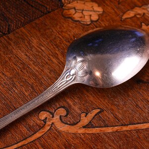 Puede incluir: Una cuchara de metal plateado con un motivo floral en relieve en la base del cuenco donde se une al mango. El mango muestra un patr&oacute;n lineal e incluye peque&ntilde;as marcas de texto estampadas cerca del elemento floral decorativo.