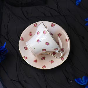 Może przedstawiać: Biała porcelanowa filiżanka do herbaty ze spodkiem, ozdobiona powtarzającym się wzorem małych r&oacute;żowych r&oacute;ż z zielonymi liśćmi. Filiżanka posiada zakrzywione ucho, a spodek ma falisty brzeg, oba wykończone cienką złotą obw&oacute;dką na krawędziach.