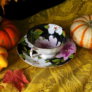 May include: A black teacup and saucer set with a floral design featuring white and pink flowers and green leaves. The teacup and saucer are on a yellow lace tablecloth, with pumpkins in the background.